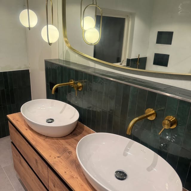 Zwei weisse Aufsatzwaschbecken auf einer Holzkonsole vor dunkelgrünen Platten mit goldfarbenen Armaturen